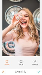 Tutorial de edição da foto de uma mulher segurando um donuts em frente ao olho e sorrindo usando a ferramenta Clarear do AirBrush