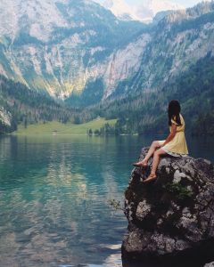 Foto de uma mulher sentada em uma rocha com uma paisagem com montanhas e lago ao fundo. 