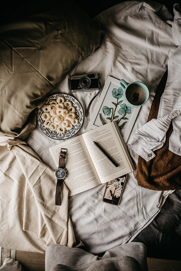Foto de vários objetos espalhados na cama. Relógio, máquina fotográfica, livro, caneta, celular e café. 