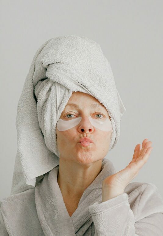 Foto de uma senhora com toalha enrolada no cabelo, mandando beijinho para a câmera fazendo skin care. 