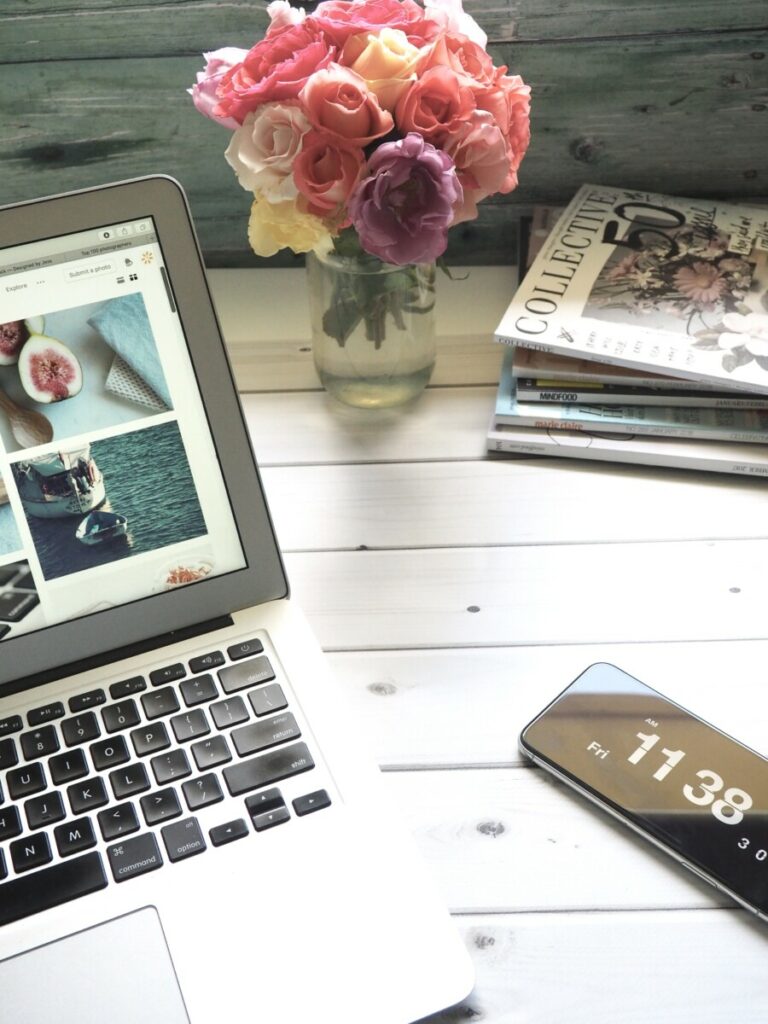 Uma imagem de um notebook aberto em um site de imagens, revistas e um jarro de flores junto com um celular em cima de uma mesa