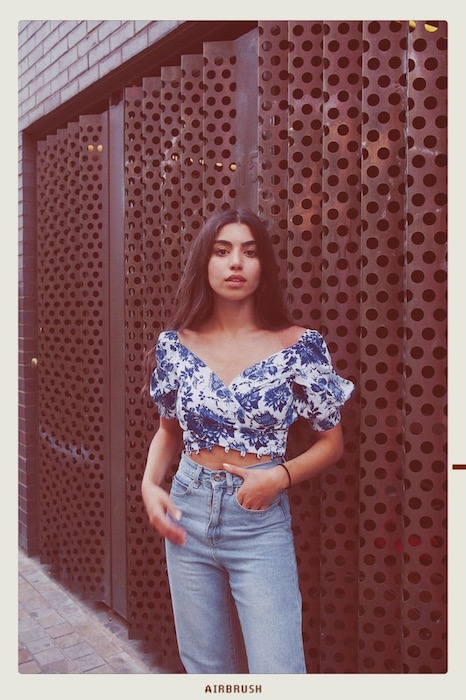 faded photo Kim Kardashian lookalike in blue jeans and floral top standing in front of metal wall