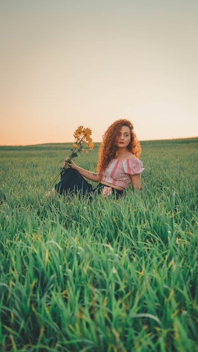 Picture of a ginger woman holding yellow flowers sitting in grass at the open field