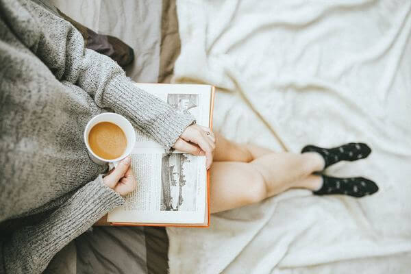 Foto tomada desde arriba de una persona con un libro apoyado sobre sus piernas y una taza de café.