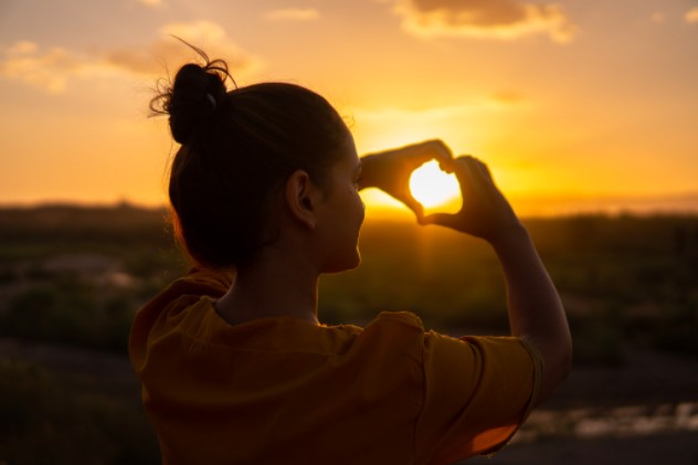 Una imagen de una niña con el pelo lacio, atada en un moño. Haciendo un corazón con sus manos, en vista de una puesta de sol en la montaña.