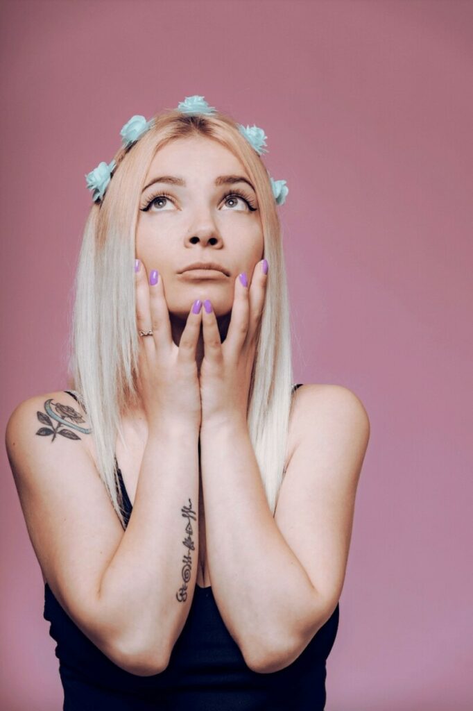 Blonde woman with blue flowers in her hair, with both hands on her face and starring up