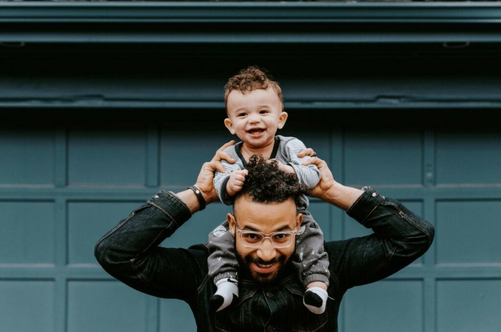 father holding his toddler son on his shoulders in front of green garage wall