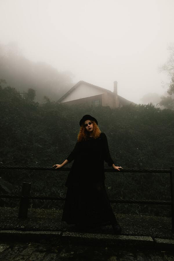 Garota usando vestido e chapéu preto com um muro de plantas ao fundo e uma casa coberta pla neblina