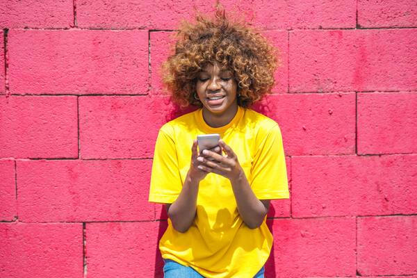 Uma garota negra de camiseta amarela segurando um celular numa parede vermelha ao fundo