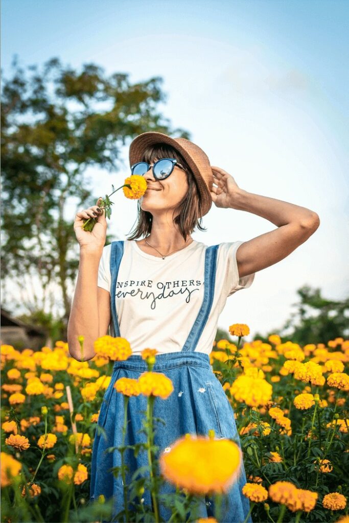 woman wearing a white tshirt and denim overalls smells a yellow flower