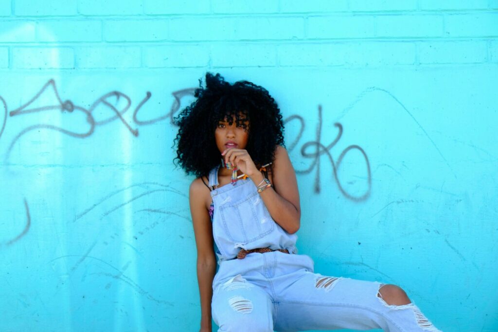 black woman wearing denim overalls sitting in front of a blue wall